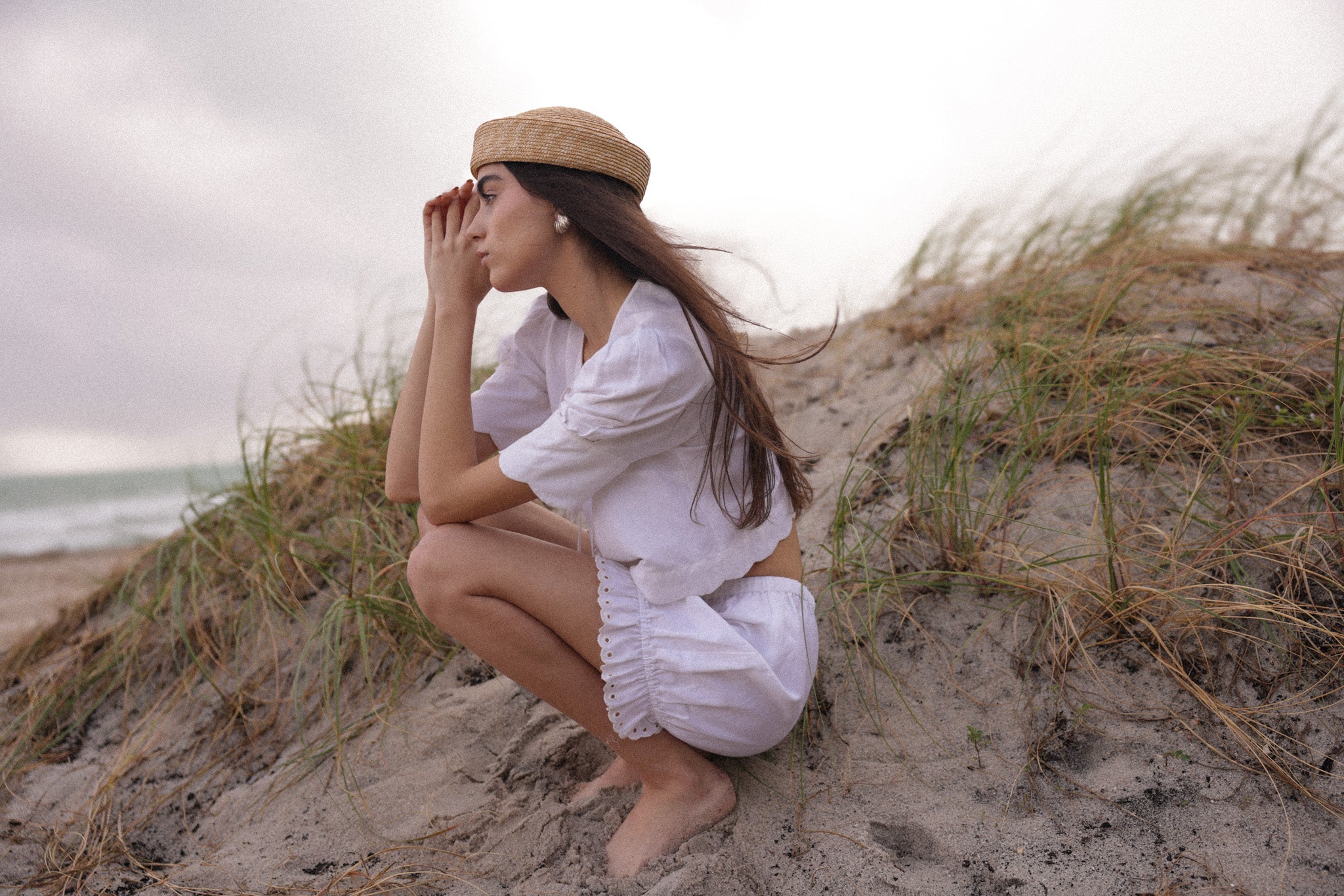Woman wearing white linen tie-front top and matching shorts sitting barefoot on sand dunes near the ocean, coastal summer outfit by Sister New York.