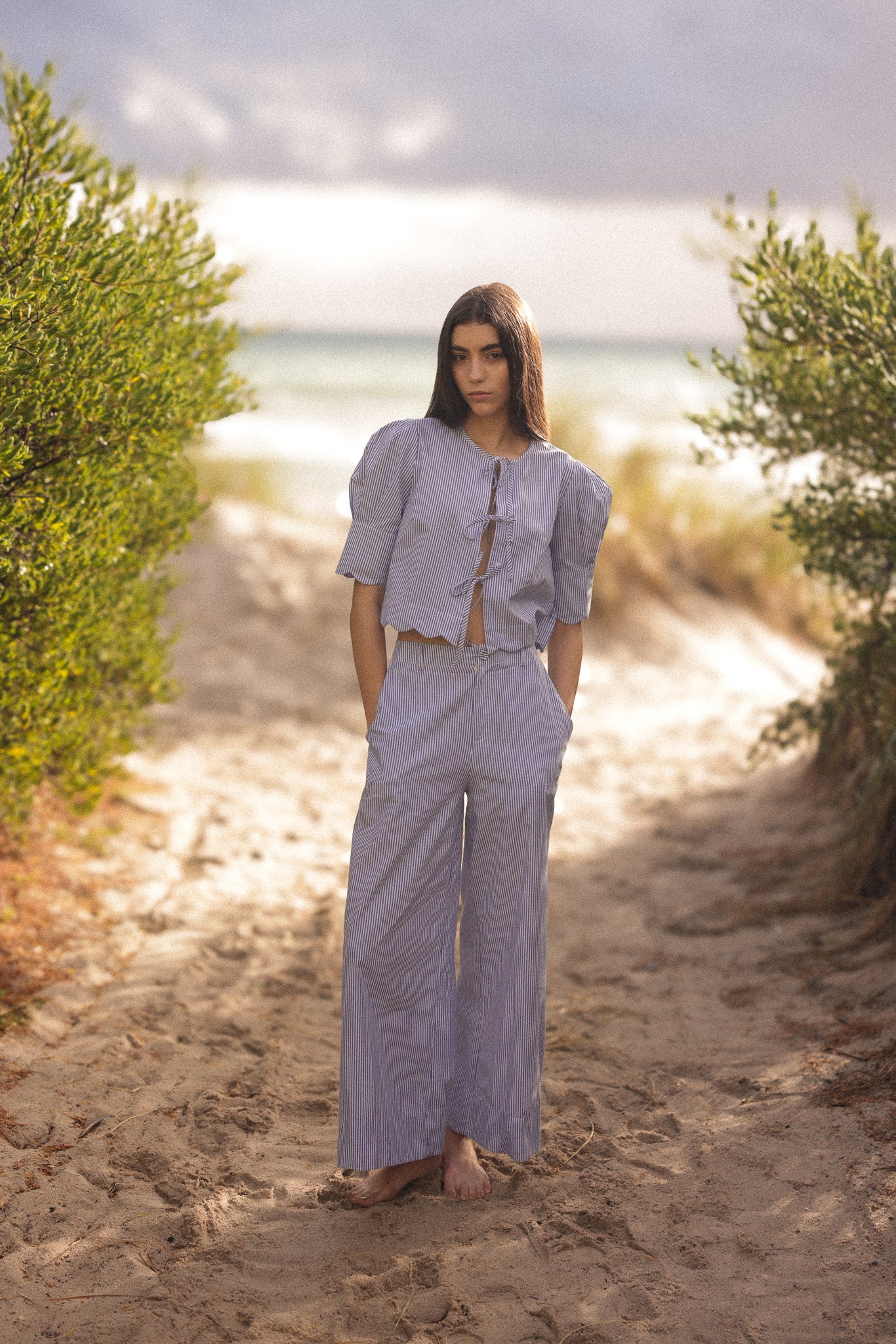Woman in a light purple outfit standing on a sandy path with greenery and water in the background