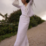 Woman in a white outfit standing on a sandy beach with greenery in the background