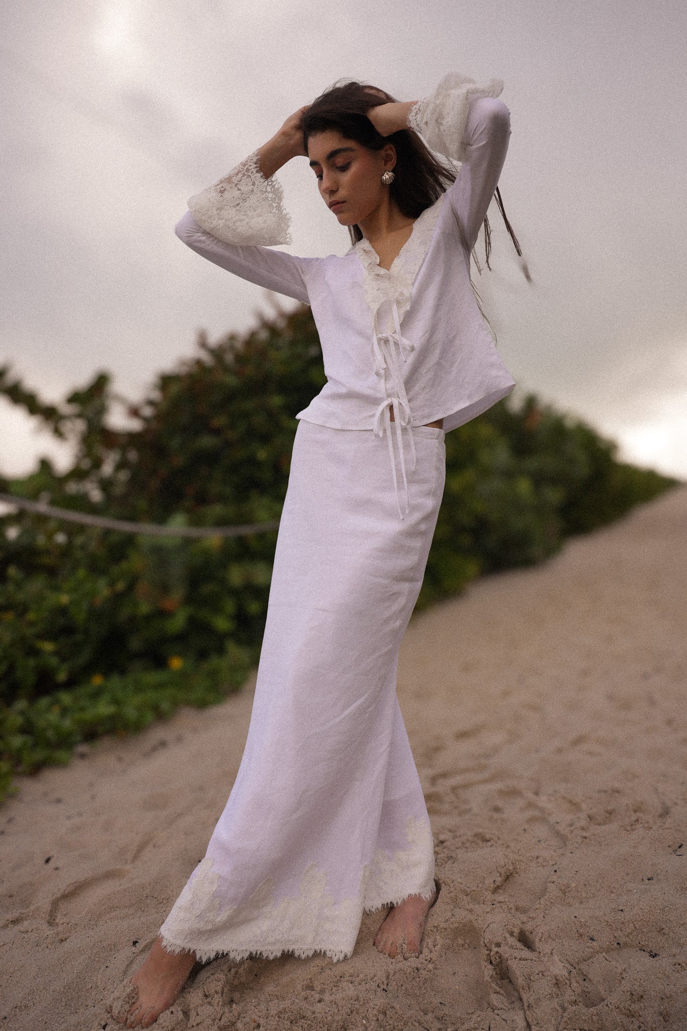 Woman in a white outfit standing on a sandy beach with greenery in the background