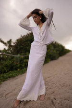 Woman in a white outfit standing on a sandy beach with greenery in the background