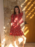 Woman in a red outfit standing against a stone wall with sunlight casting shadows.
