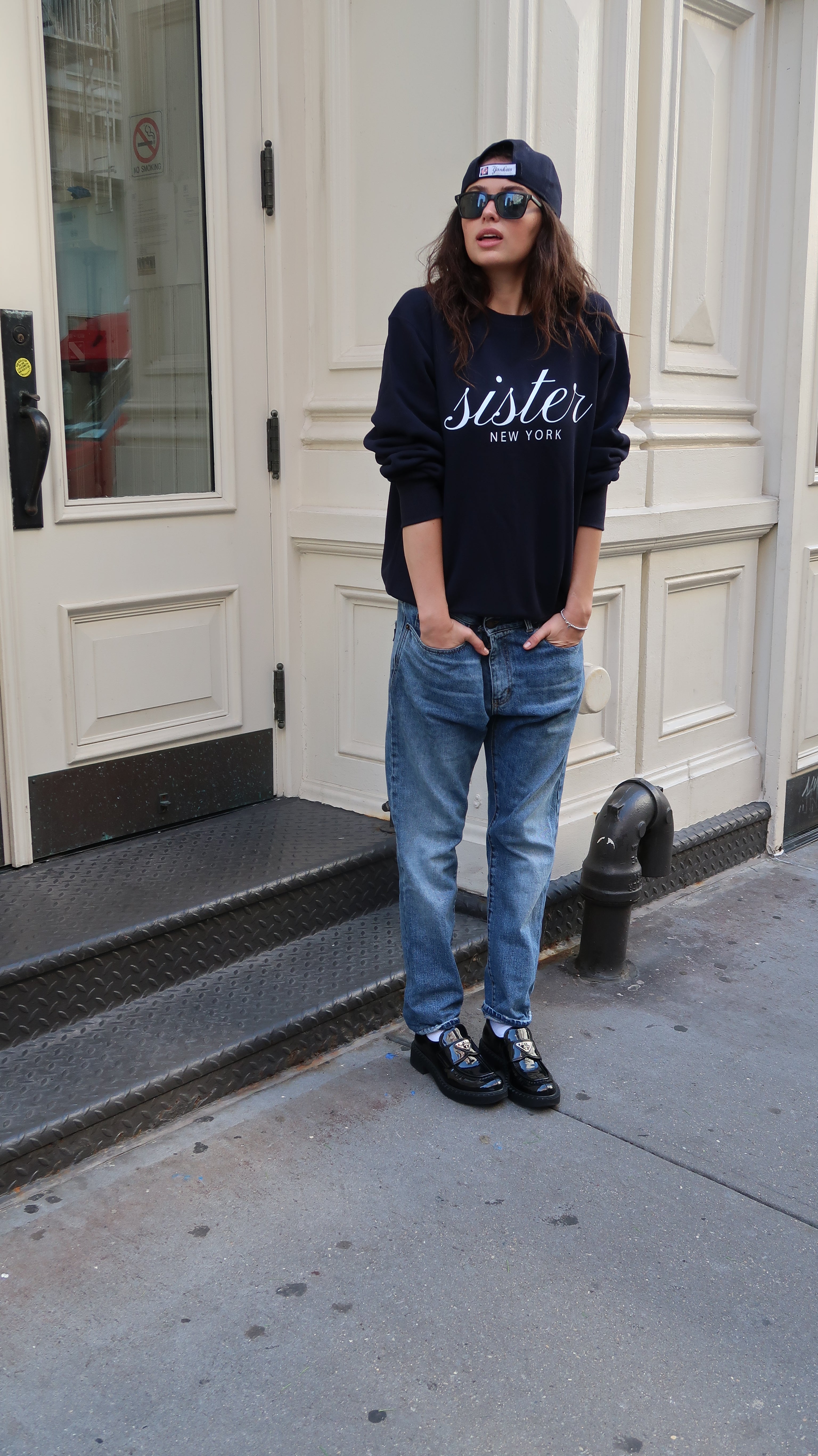 Person wearing a navy hoodie with 'Sister' text, blue jeans, and black shoes standing on a sidewalk.