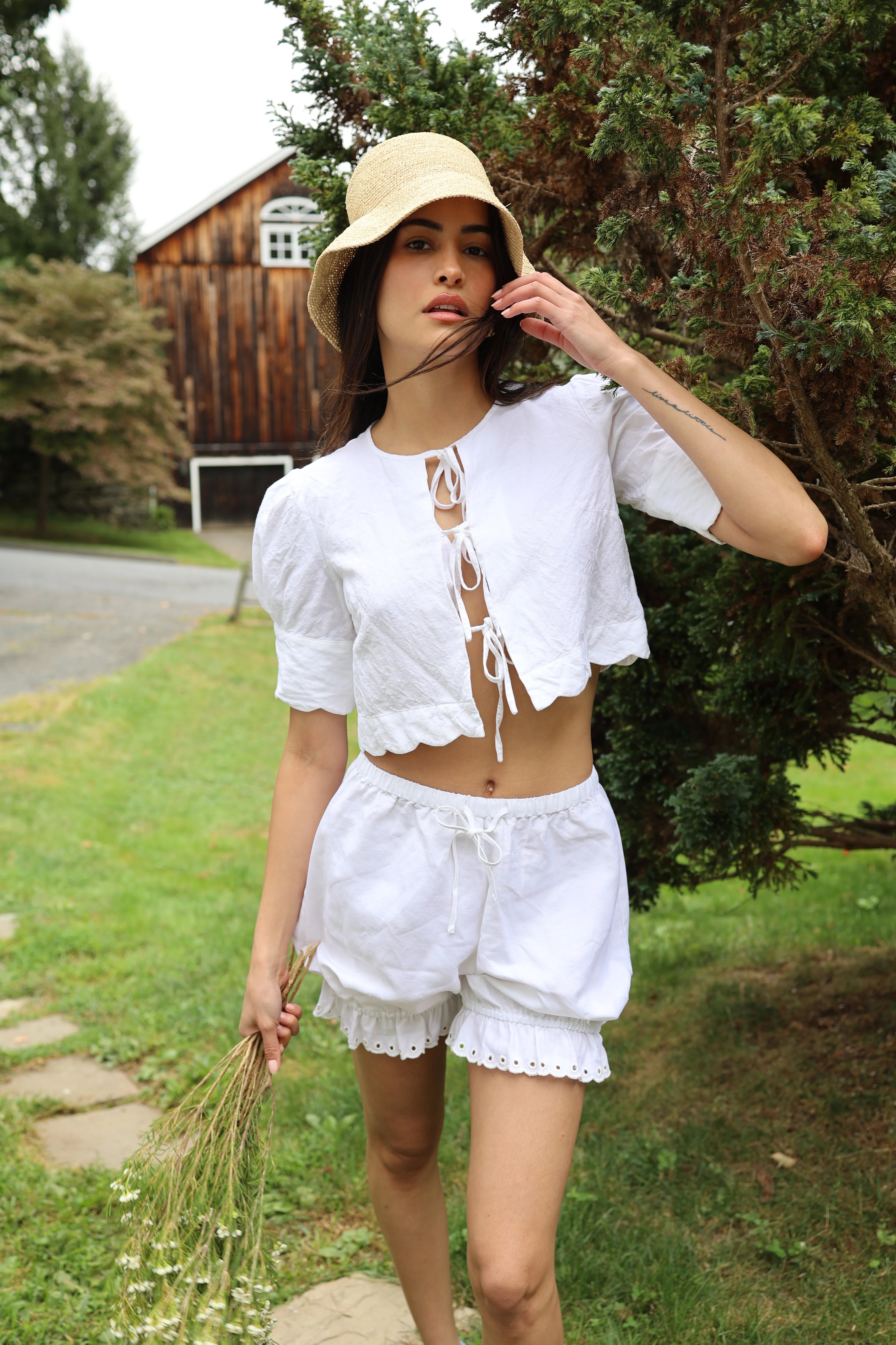 Woman in a white outfit standing outdoors with trees and a wooden building in the background
