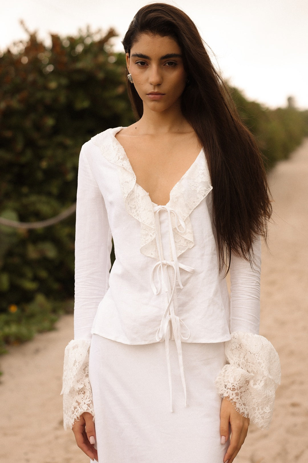 Model walking on a sandy path wearing a white lace-trimmed tie-front top and matching white skirt from Sister NY.